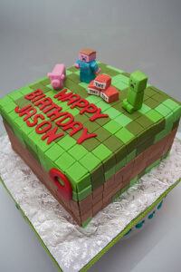 Square Minecraft-themed birthday cake with fondant pig, Creeper, TNT blocks, and “Happy Birthday Jason” lettering on a pixelated green and brown surface.