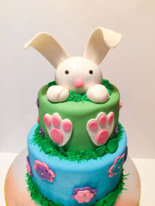 Two-tier Easter bunny cake with a fondant bunny peeking over the top, featuring large ears, paws, and colorful fondant flowers on blue and green tiers.