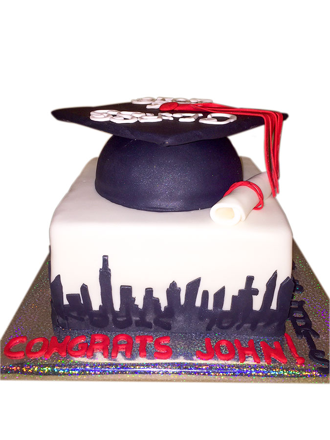 Graduation-themed fondant cake featuring a black mortarboard cap, diploma scroll, and a city skyline design with red 'Congrats John!' lettering on a glittery cake board.