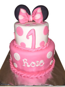 Two-tier pink and white Minnie Mouse fondant cake with polka dots, a pink bow and ears topper, featuring a number 1 and personalized with the name Rose for a first birthday celebration.