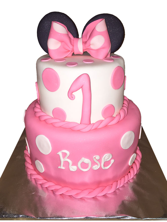 Two-tier pink and white Minnie Mouse fondant cake with polka dots, a pink bow and ears topper, featuring a number 1 and personalized with the name Rose for a first birthday celebration.