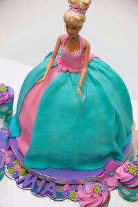 Barbie-themed birthday cake featuring a doll with a teal and pink fondant dress, designed as the cake’s centerpiece, surrounded by matching cupcakes decorated with pastel flowers and edible gems for a little girl's party.