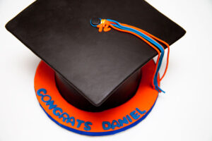 Graduation cap-shaped cake with orange and blue tassels, featuring “Congrats Daniel” text on the base.